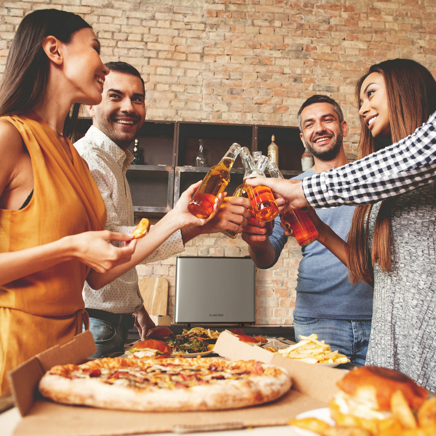 Vier Personen stoßen mit Bierflaschen an, um einen Tisch mit Pizza und Snacks herum.