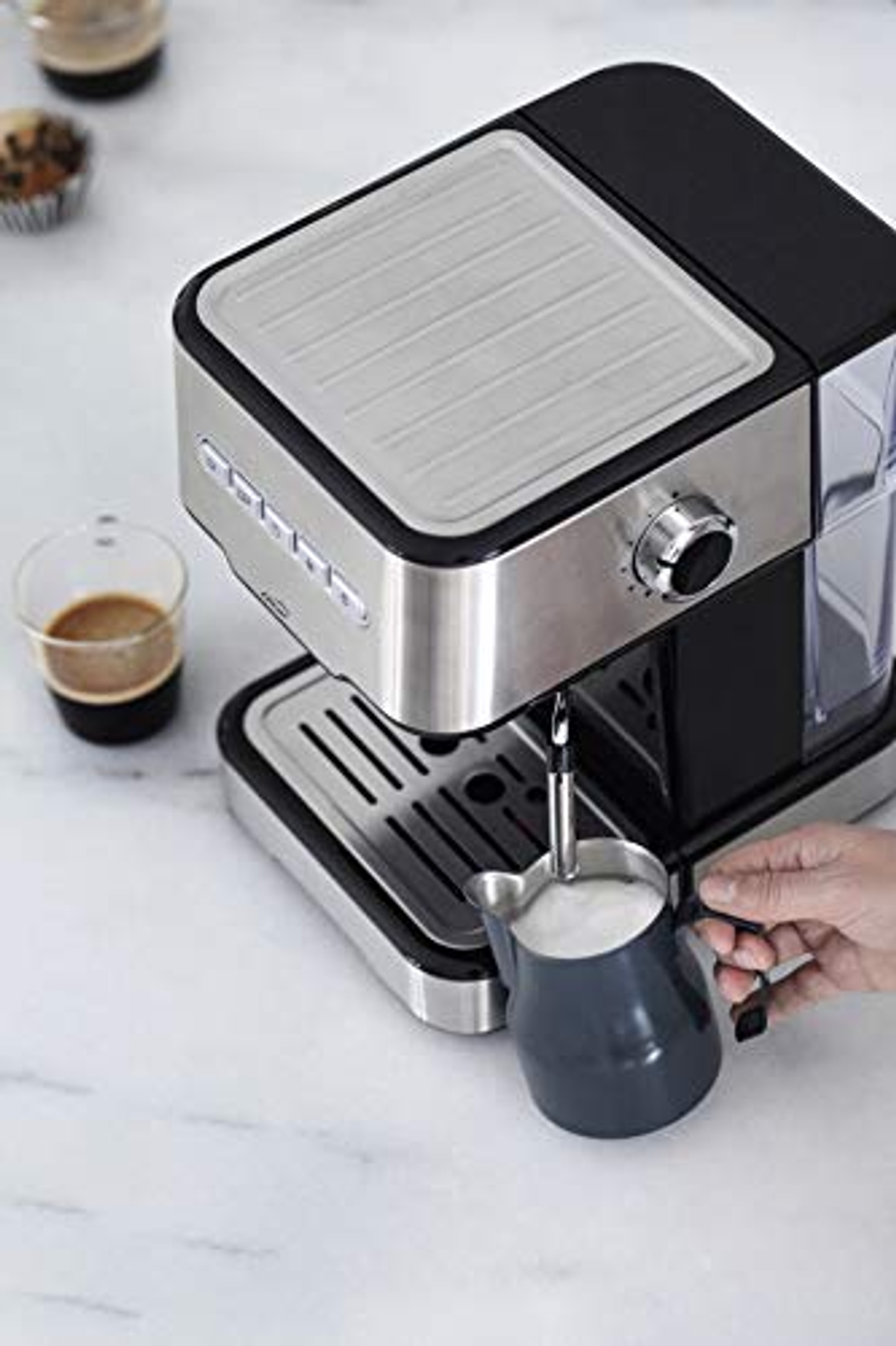 Máquina de espresso haciendo café. Una mano sostiene una jarra de leche. Encimera de mármol blanco.