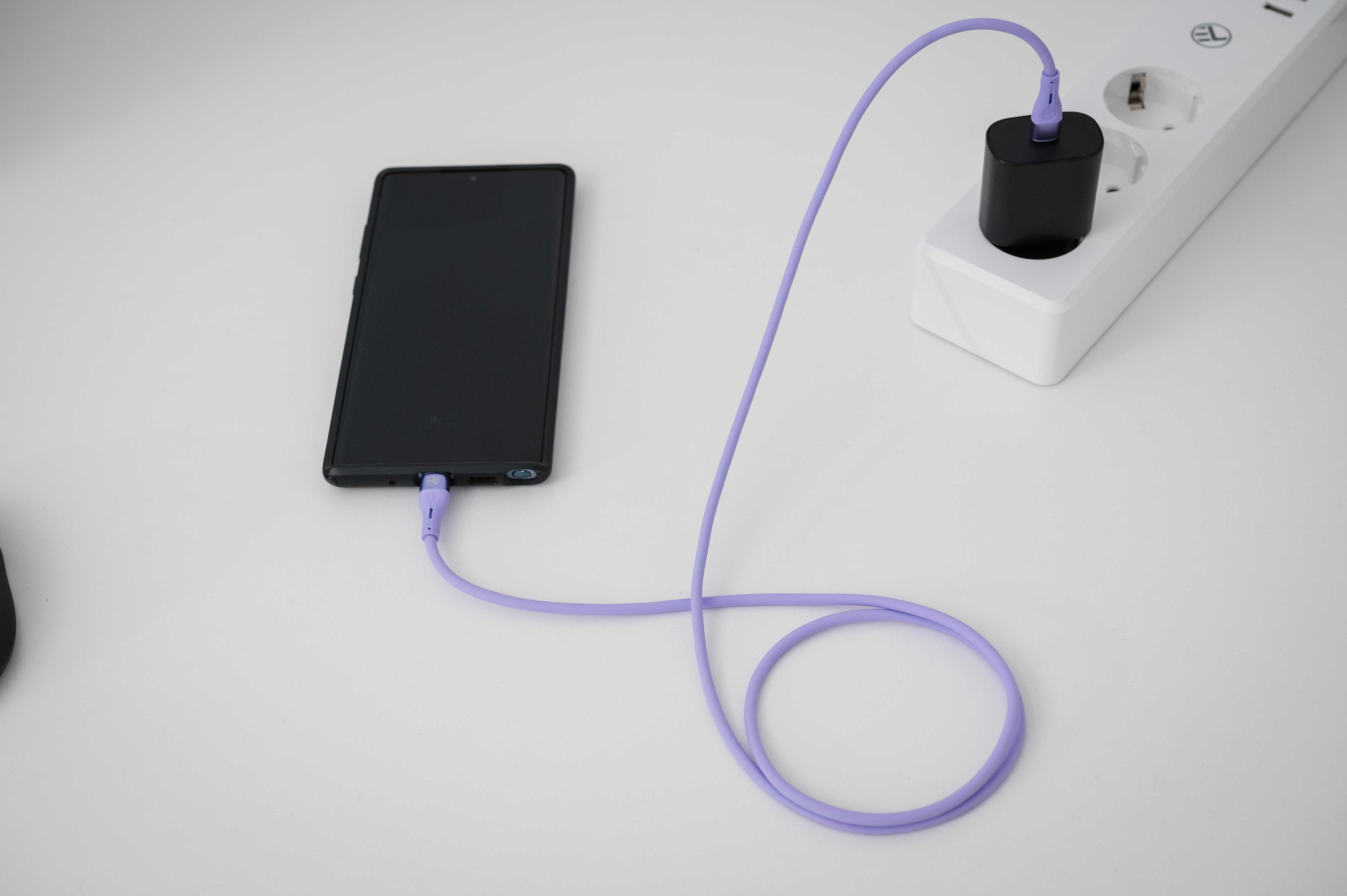 Telefon ładujący się fioletowym kablem. Ładowarka podłączona. Biała powierzchnia.