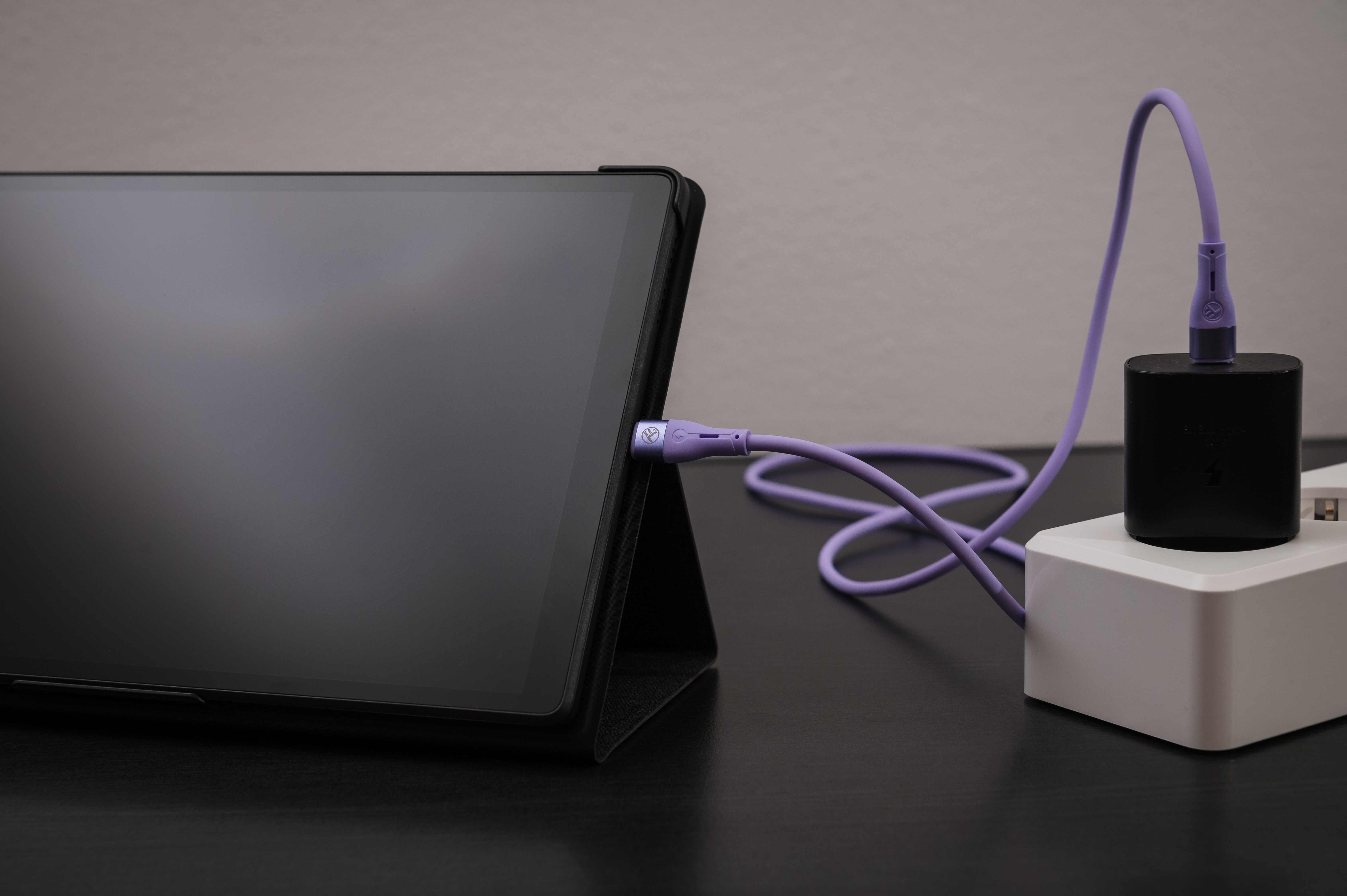 Tablet ładujący się fioletowym kablem USB-C. Czarna ładowarka w białym gniazdku.