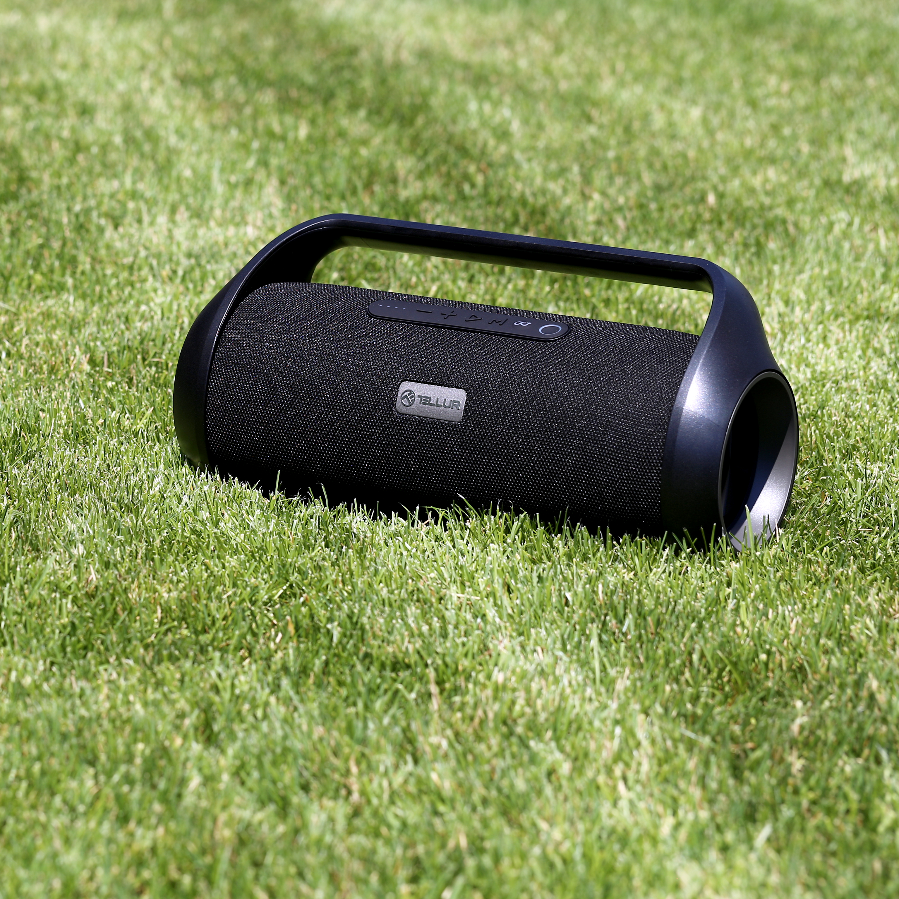Czarny głośnik przenośny na zielonej trawie. Głośnik ma uchwyt i markę 'Tellur'.