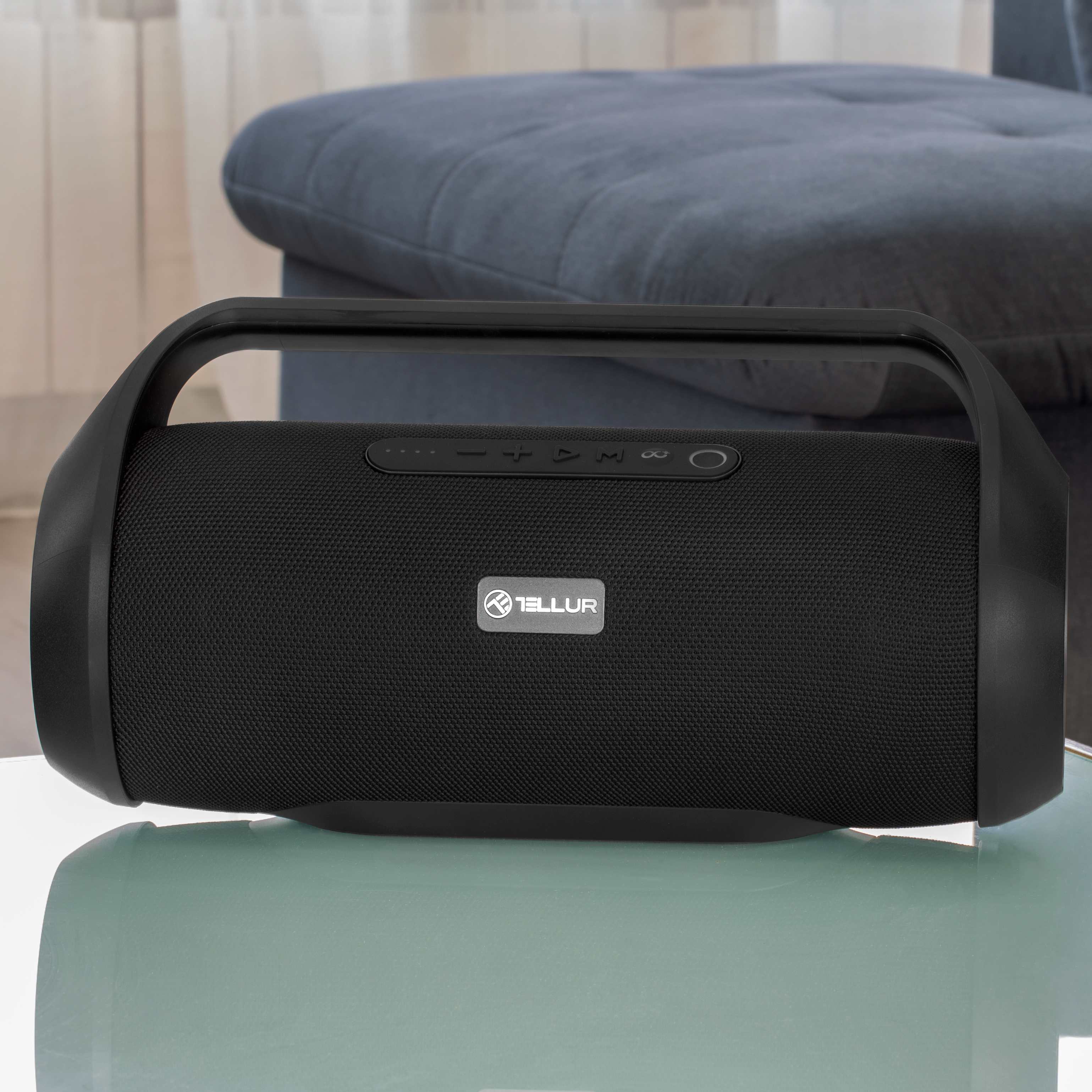 Czarny głośnik na szklanym stole. Szara kanapa w tle. Uchwyt na górze.