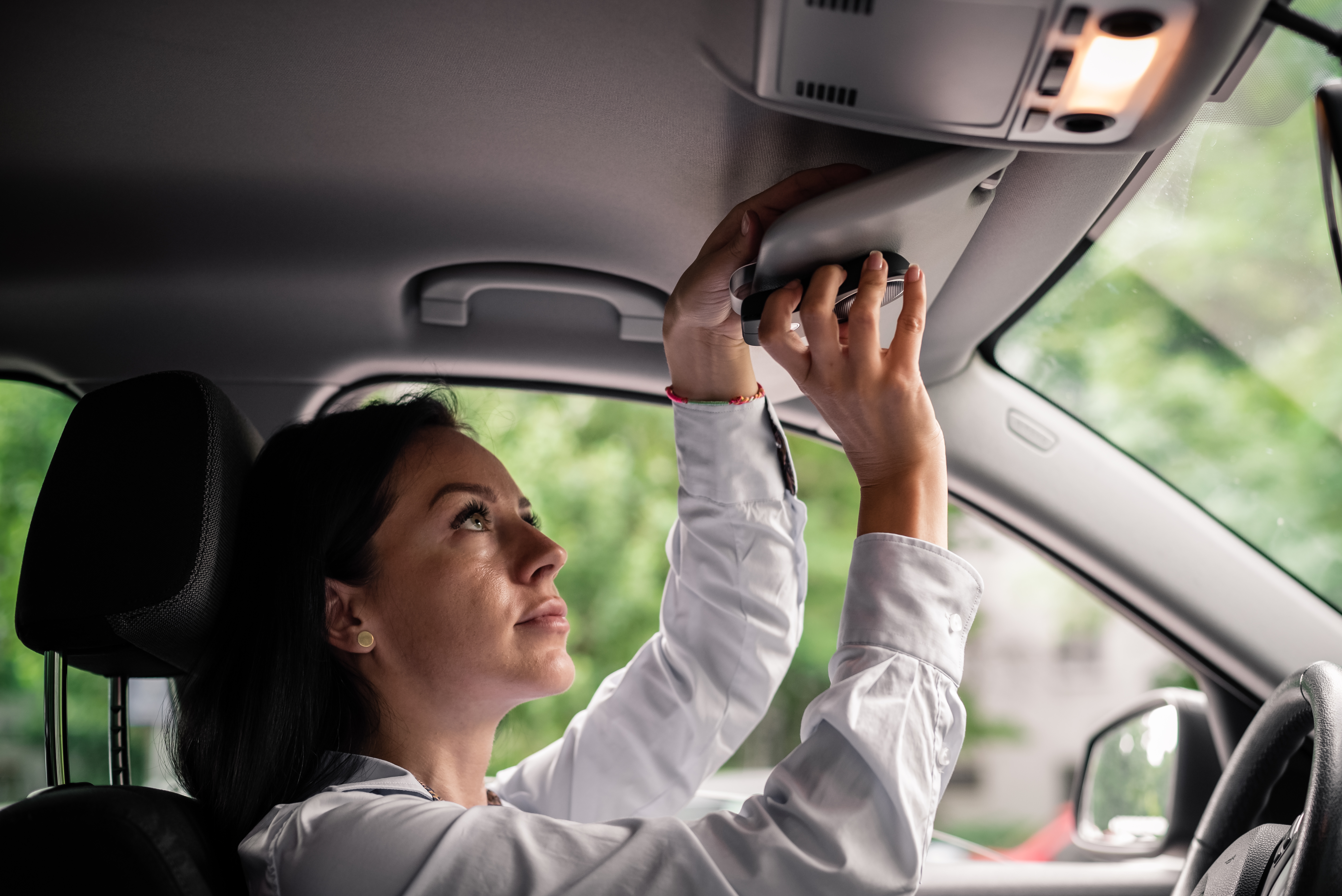 Mujer con camisa blanca dentro de un coche, ajustando el parasol. Fondo verde exterior.