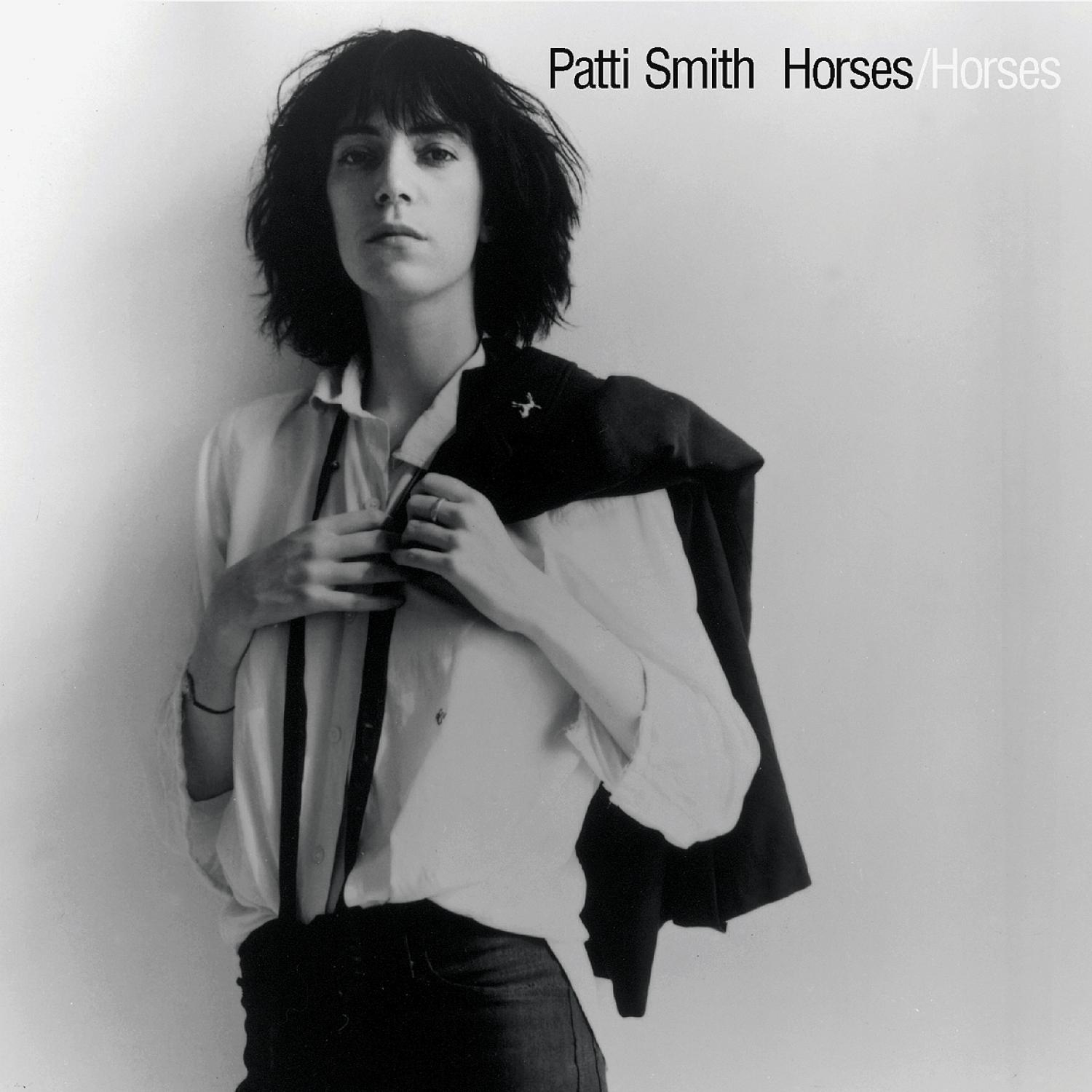 Patti Smith in einem Schwarz-Weiß-Foto. Sie trägt ein weißes Hemd und Jeans und hält eine Jacke.