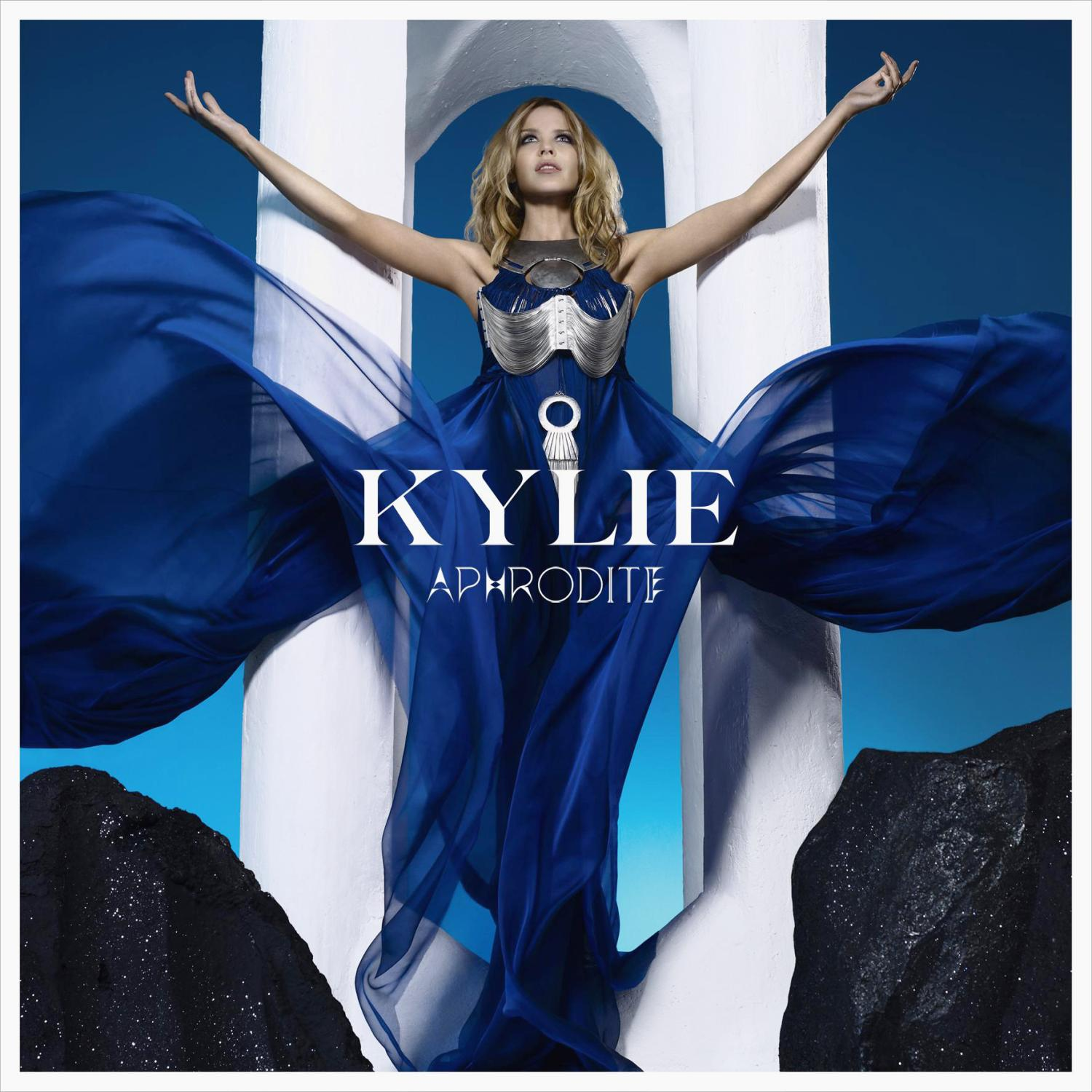 Kylie in blauem Kleid mit ausgestreckten Armen, Titel 'Kylie Aphrodite' in Weiß. Weißer Bogen und schwarze Felsen im Hintergrund.
