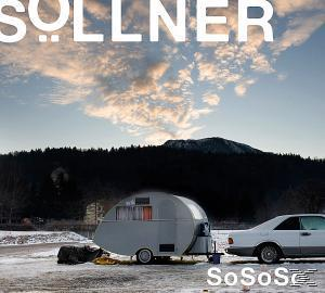 Ein silberner Wohnwagen parkt neben einem weißen Auto. Ein Berg und ein bewölkter Himmel im Hintergrund.