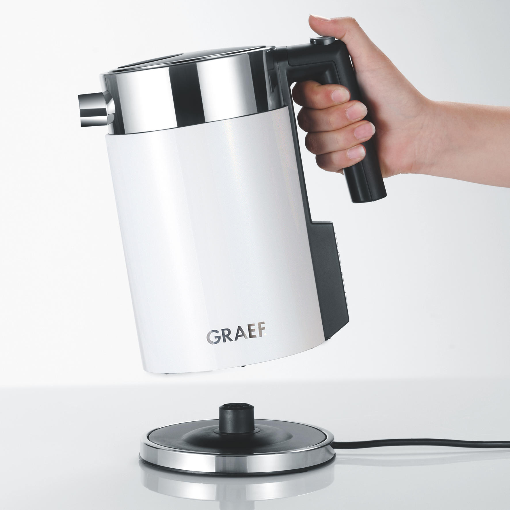 Ein weißer Wasserkocher, der von einer Hand gehalten wird. Der Wasserkocher steht auf einem Sockel. Das Wort GRAEF ist sichtbar.