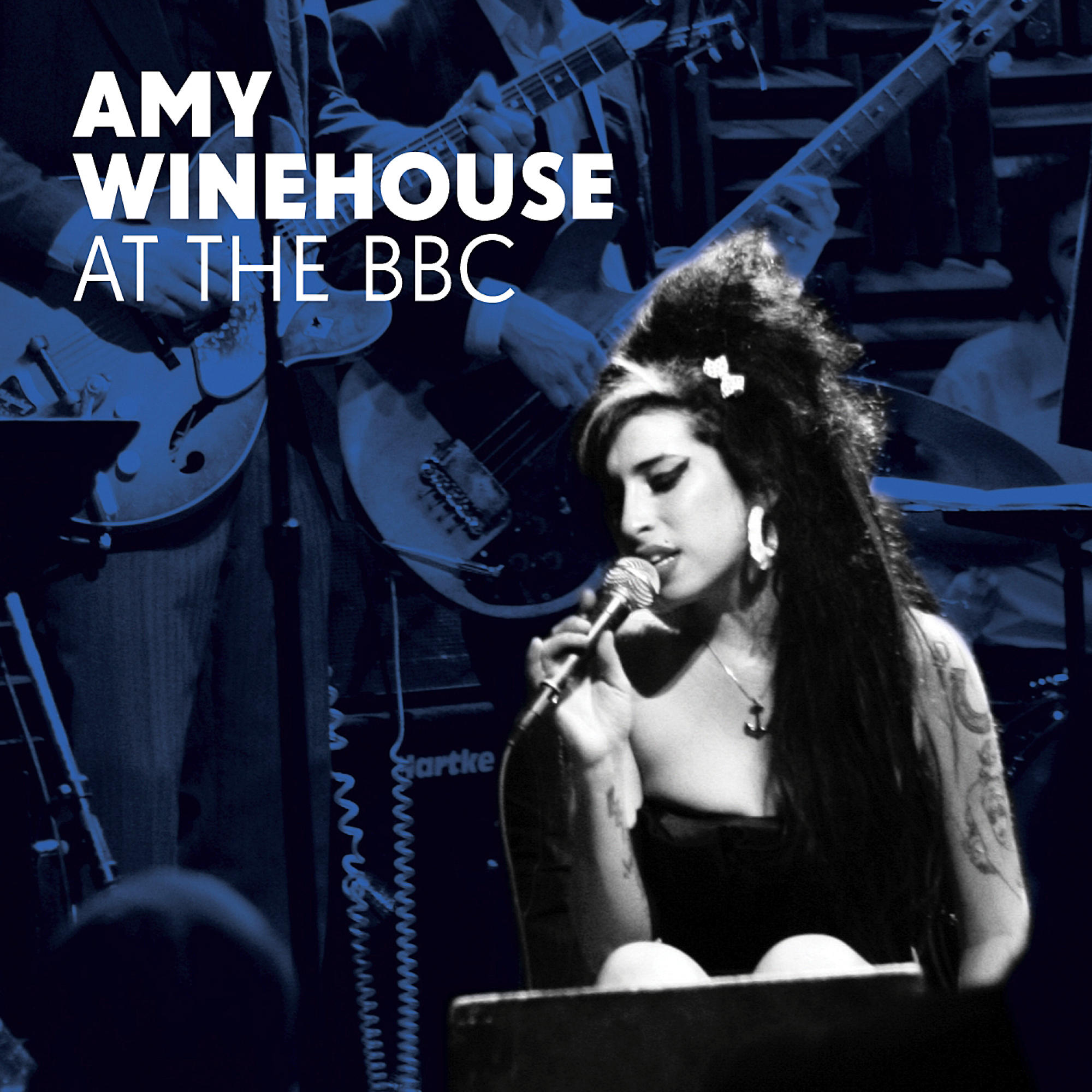 Amy Winehouse singt auf der Bühne mit Mikrofon. Blautöniges Bild mit Bandmitgliedern im Hintergrund.