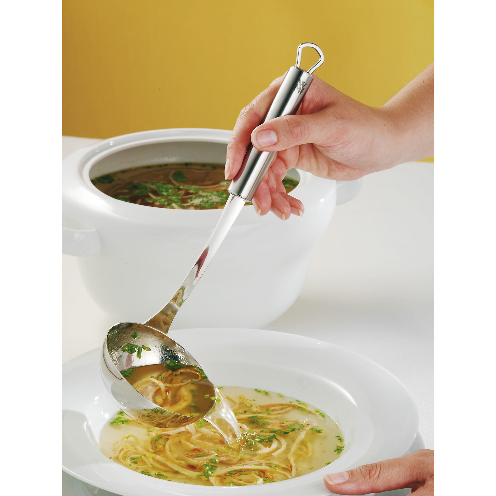 Person gießt Suppe mit einer silbernen Schöpfkelle in eine weiße Schüssel. Gelber Hintergrund.
