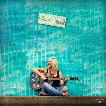 Eine Frau sitzt und spielt Gitarre vor einem blauen Wandbild mit einem Schild mit der Aufschrift 'Hong Kong'.