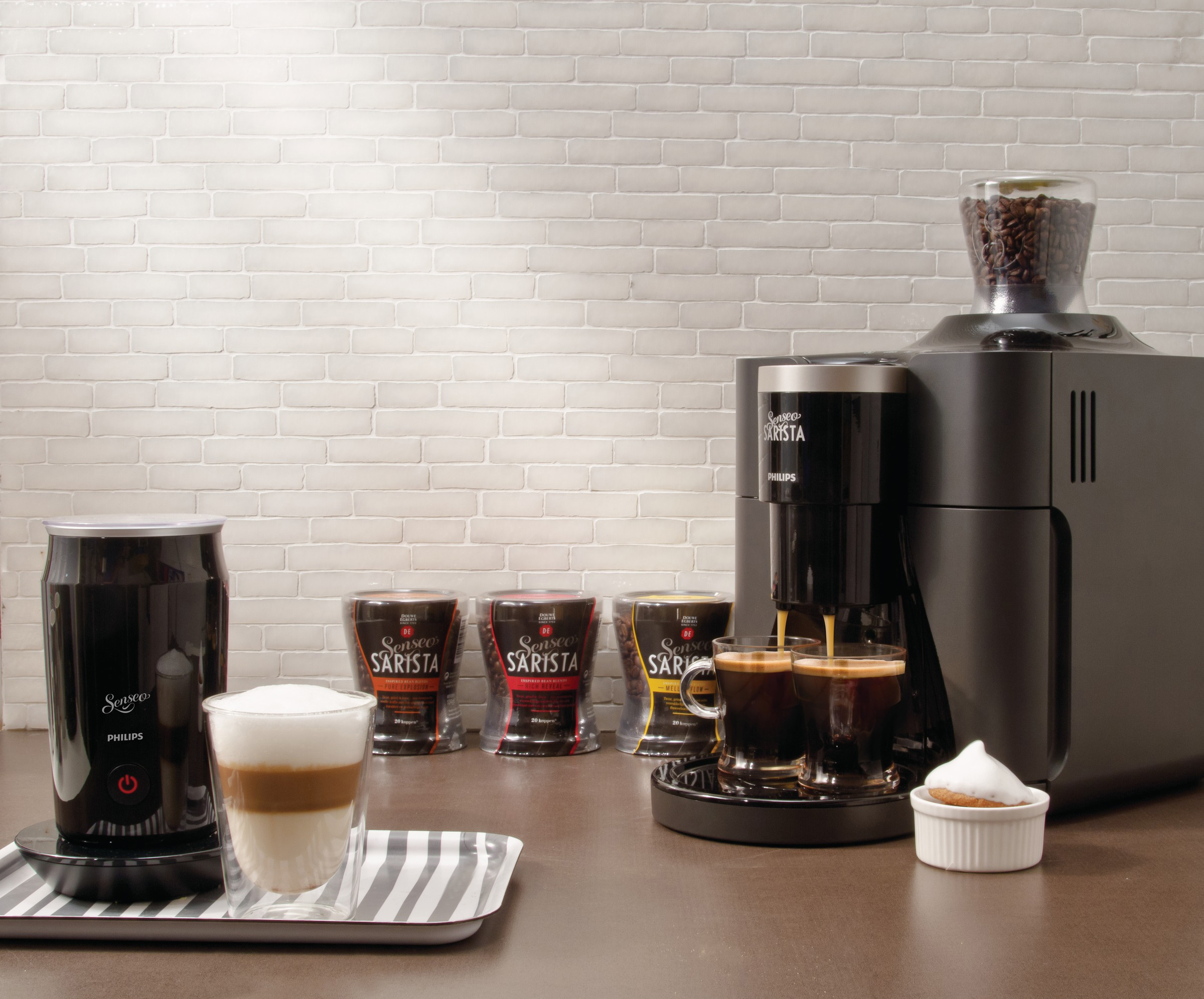 Eine Kaffeemaschine mit Latte, ein Kaffeebohnenbehälter und eine weiße Backsteinmauer im Hintergrund.