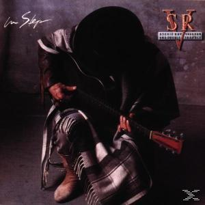 Stevie Ray Vaughan spielt Gitarre. Er trägt einen Hut und ein dunkles Outfit. Der Hintergrund ist dunkel.