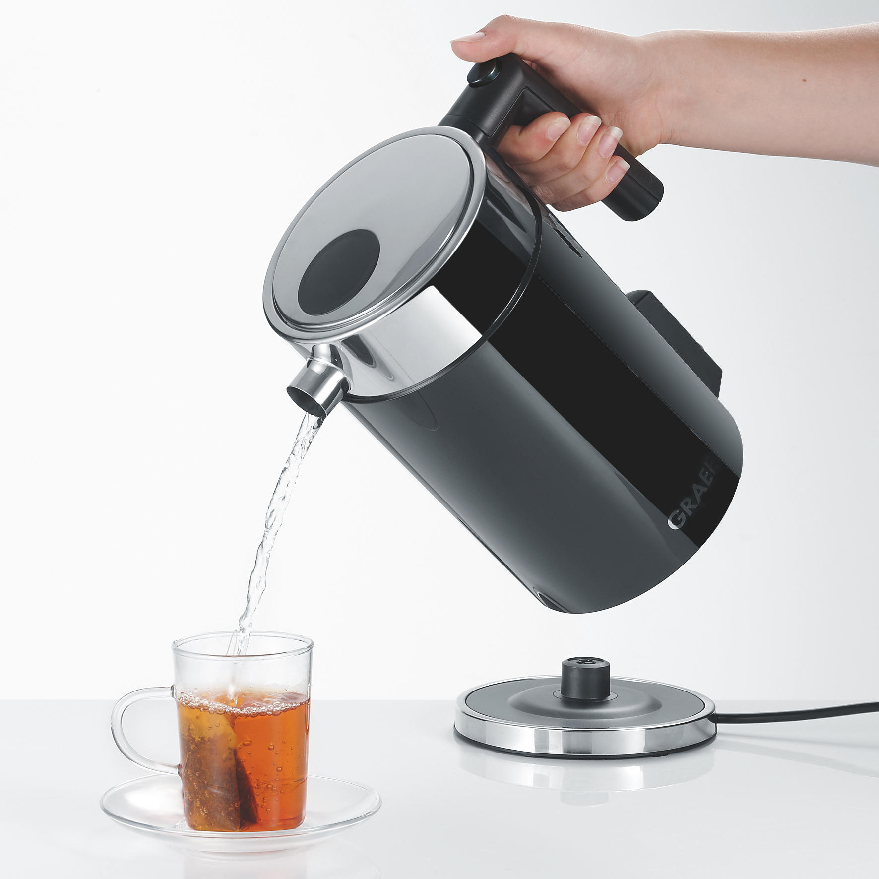 Hand gießt Wasser aus einem schwarzen Wasserkocher in eine Glastasse mit Teebeutel. Weißer Hintergrund.