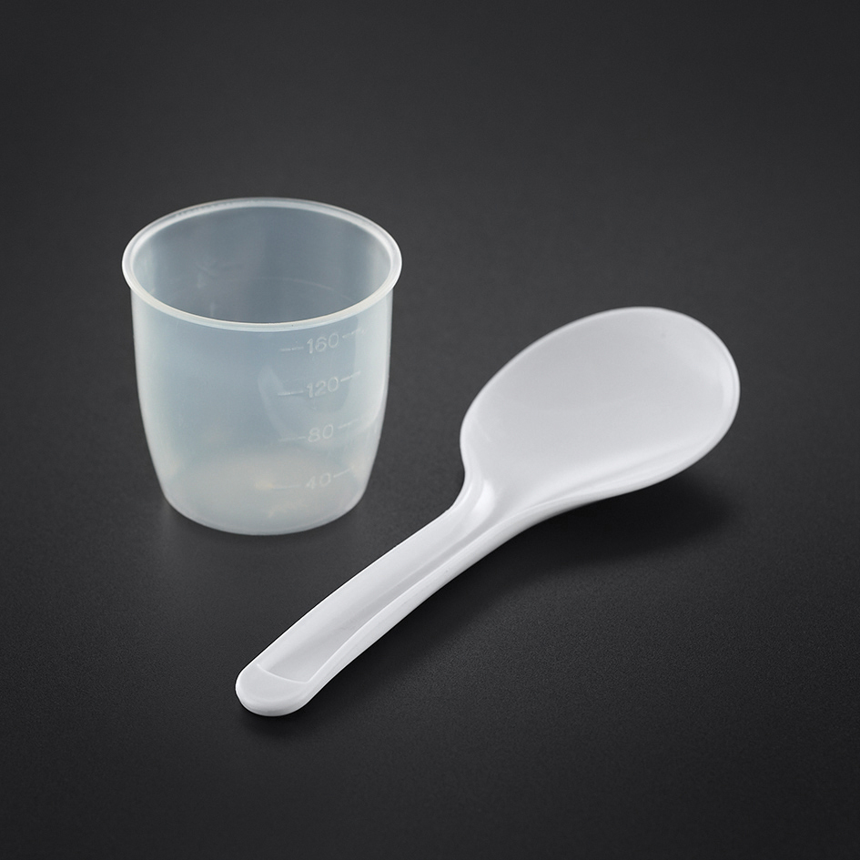 Een maatbeker en een witte lepel op een zwart oppervlak. De beker heeft meetmarkeringen.