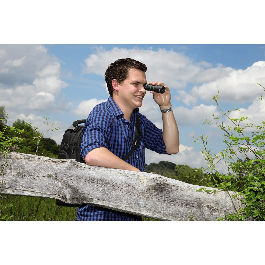 Un uomo con un binocolo guarda qualcosa. Indossa una camicia a quadri blu e uno zaino.