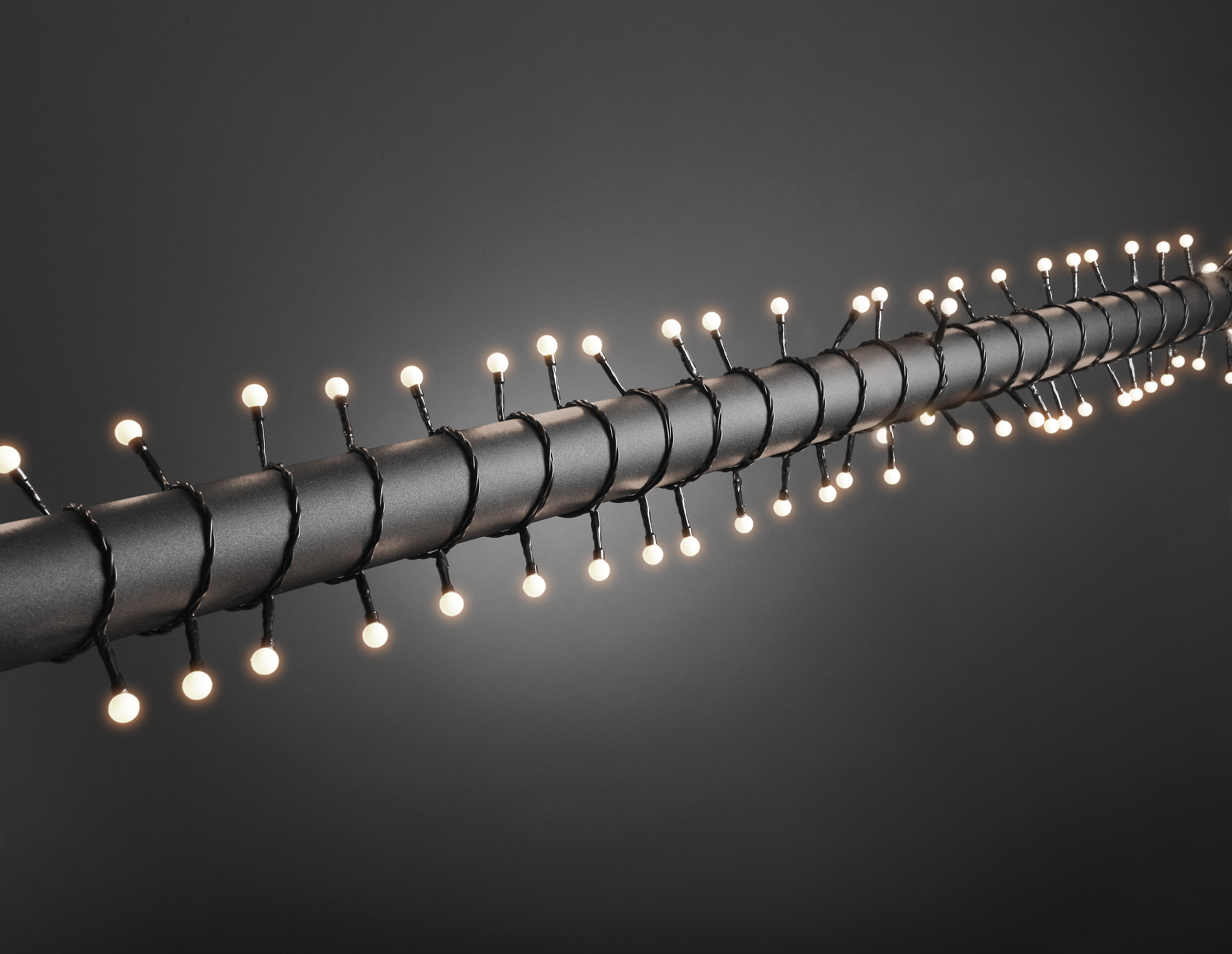 Eine Lichterkette mit kleinen weißen Glühbirnen ist um ein dunkelgraues Metallrohr vor grauem Hintergrund gewickelt.