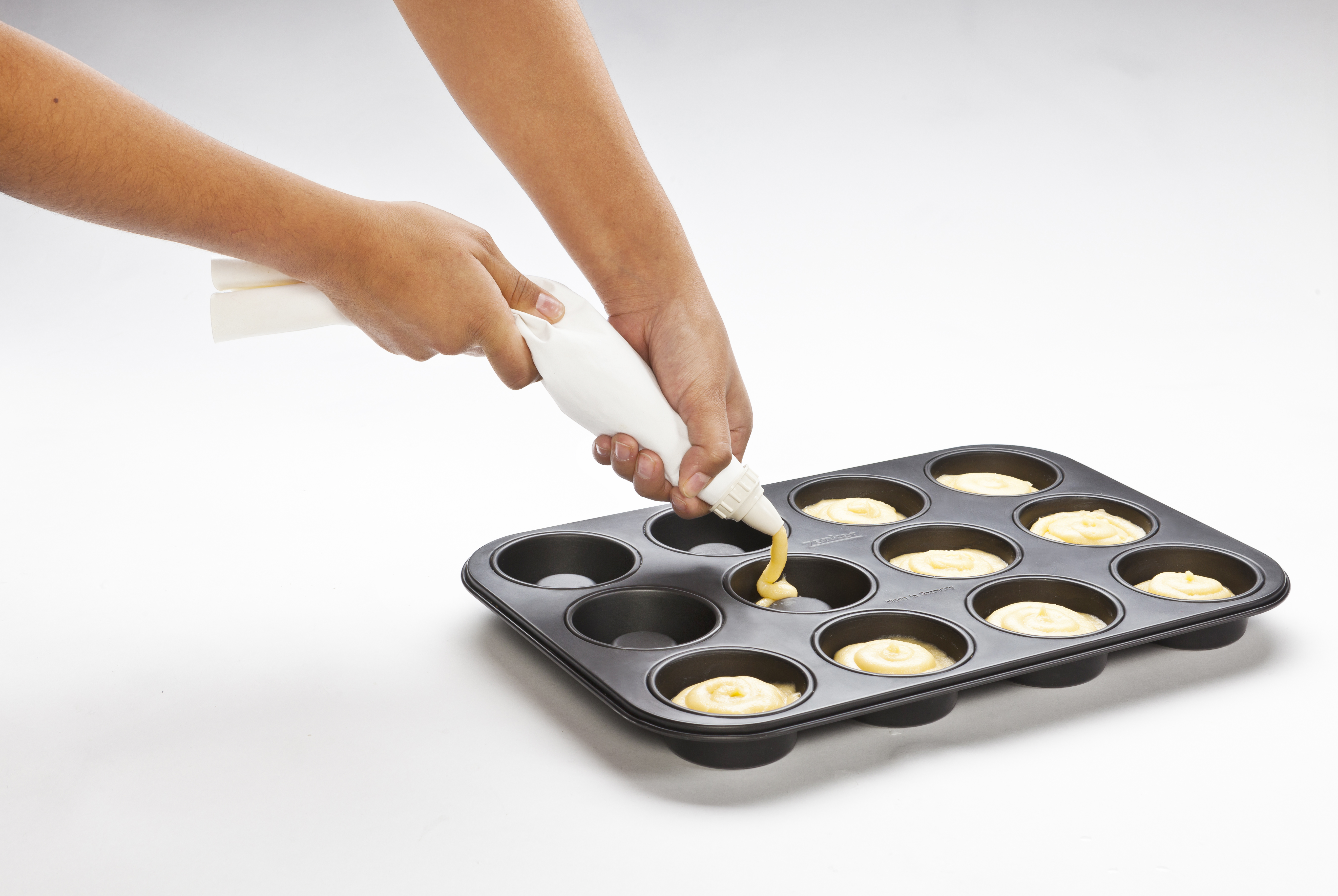 Hände füllen eine Muffinform mit Teig aus einem Spritzbeutel. Der Hintergrund ist weiß.
