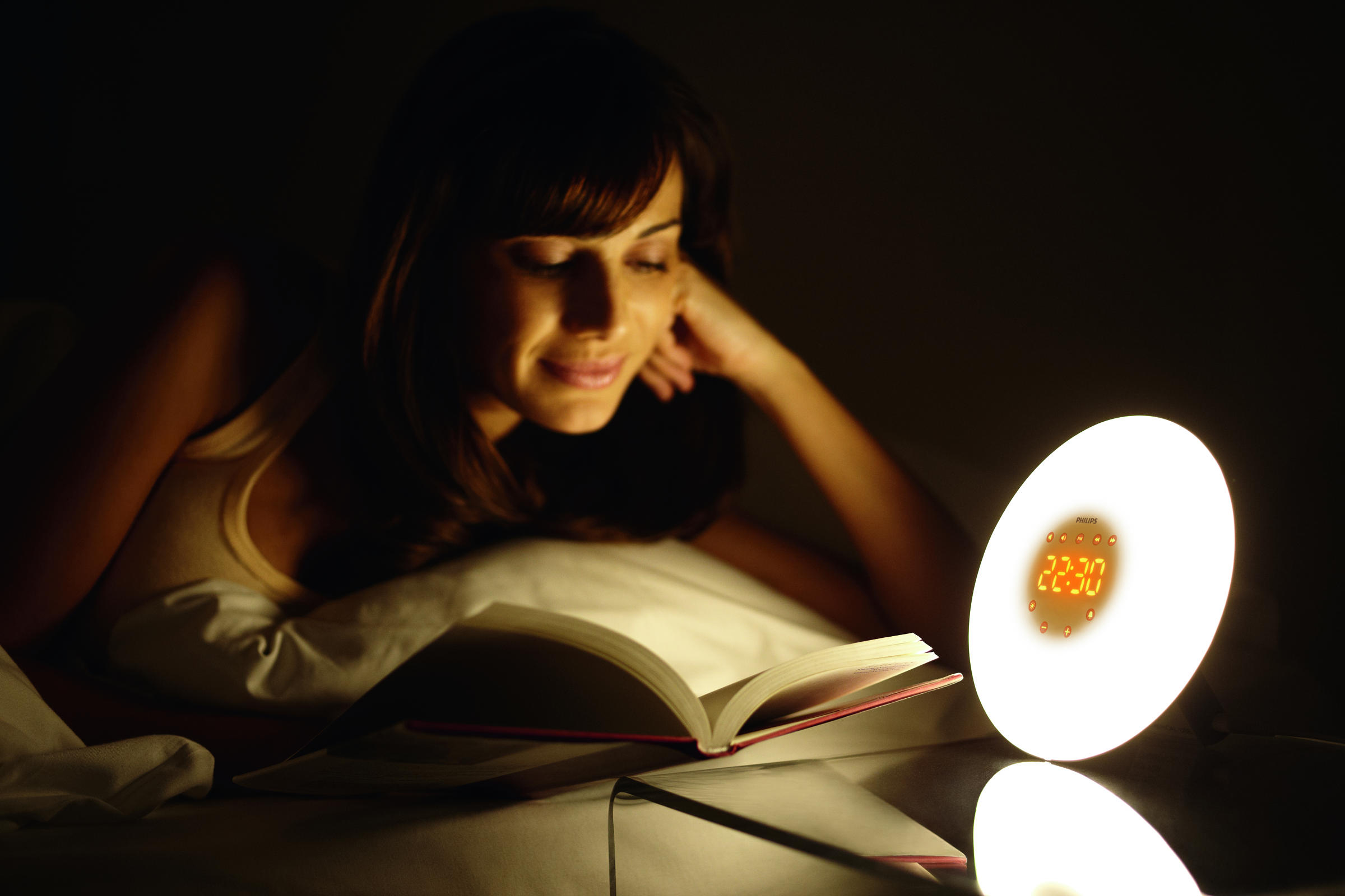 Frau liest nachts im Bett, beleuchtet von einer kreisförmigen, leuchtenden Nachttischlampe.