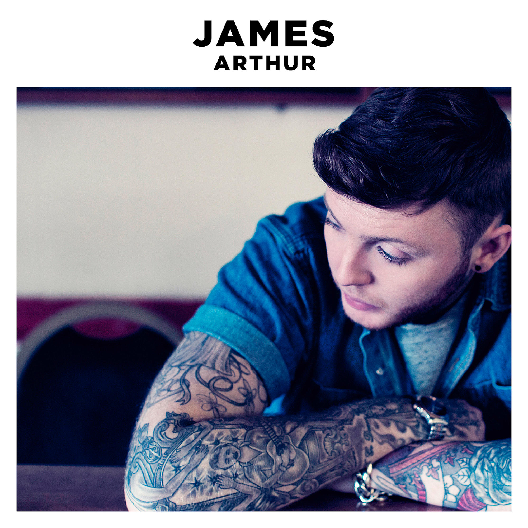 James Arthur blickt nach unten, mit Tätowierungen auf den Armen und einer Uhr.
