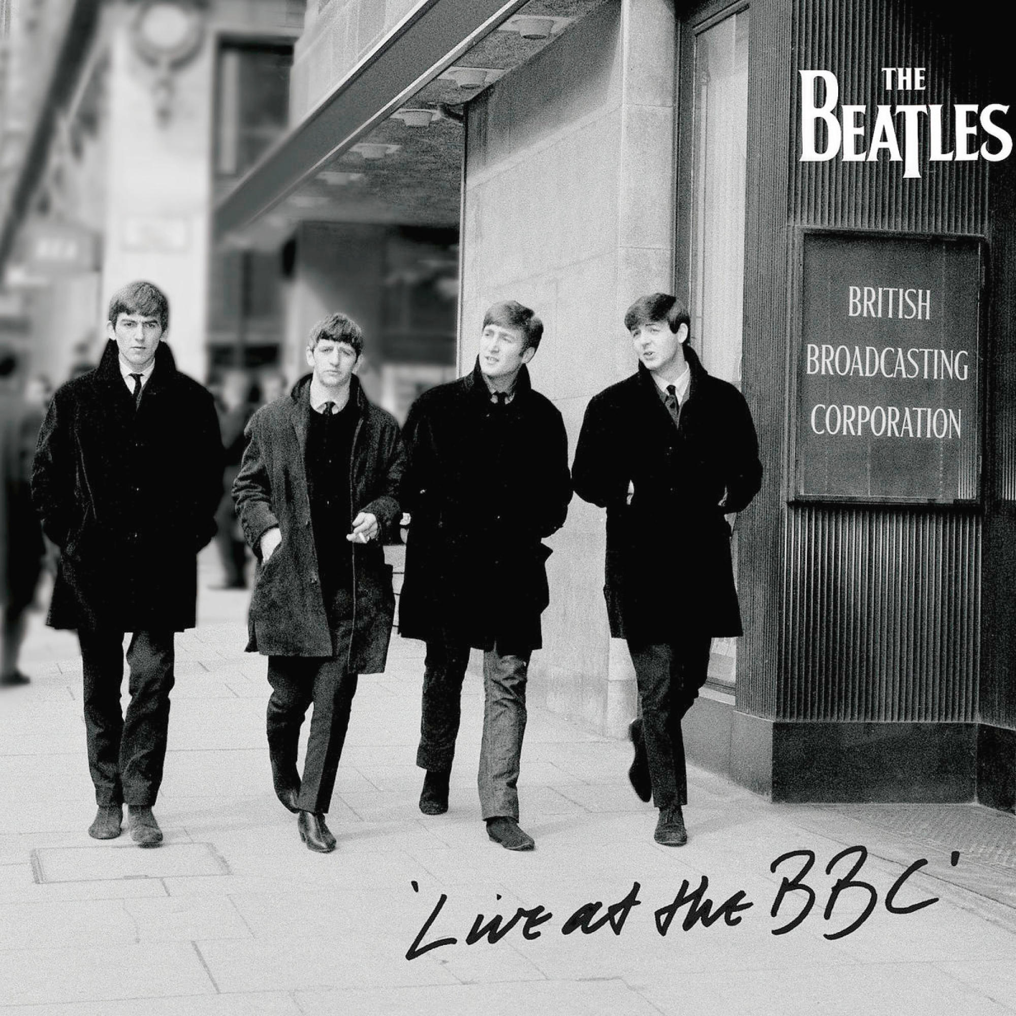 The Beatles gehen in schwarzen Mänteln auf einer Straße vor einem Gebäude.