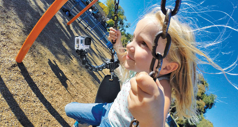 Mädchen auf einer Schaukel, hält die Ketten. GoPro-Kamera befestigt. Blauer Himmel und Spielplatz sichtbar.