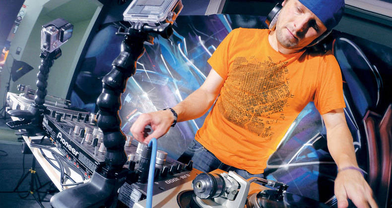 Ein DJ in einem orangefarbenen Shirt mischt Musik, mit Kameras und Graffiti-Hintergrund.