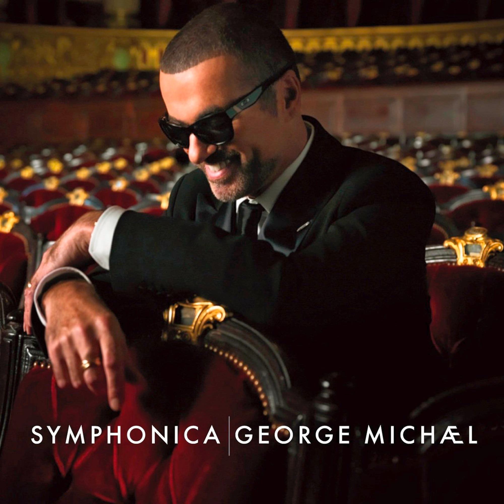 George Michael en costume et lunettes de soleil, assis dans un fauteuil en velours rouge.