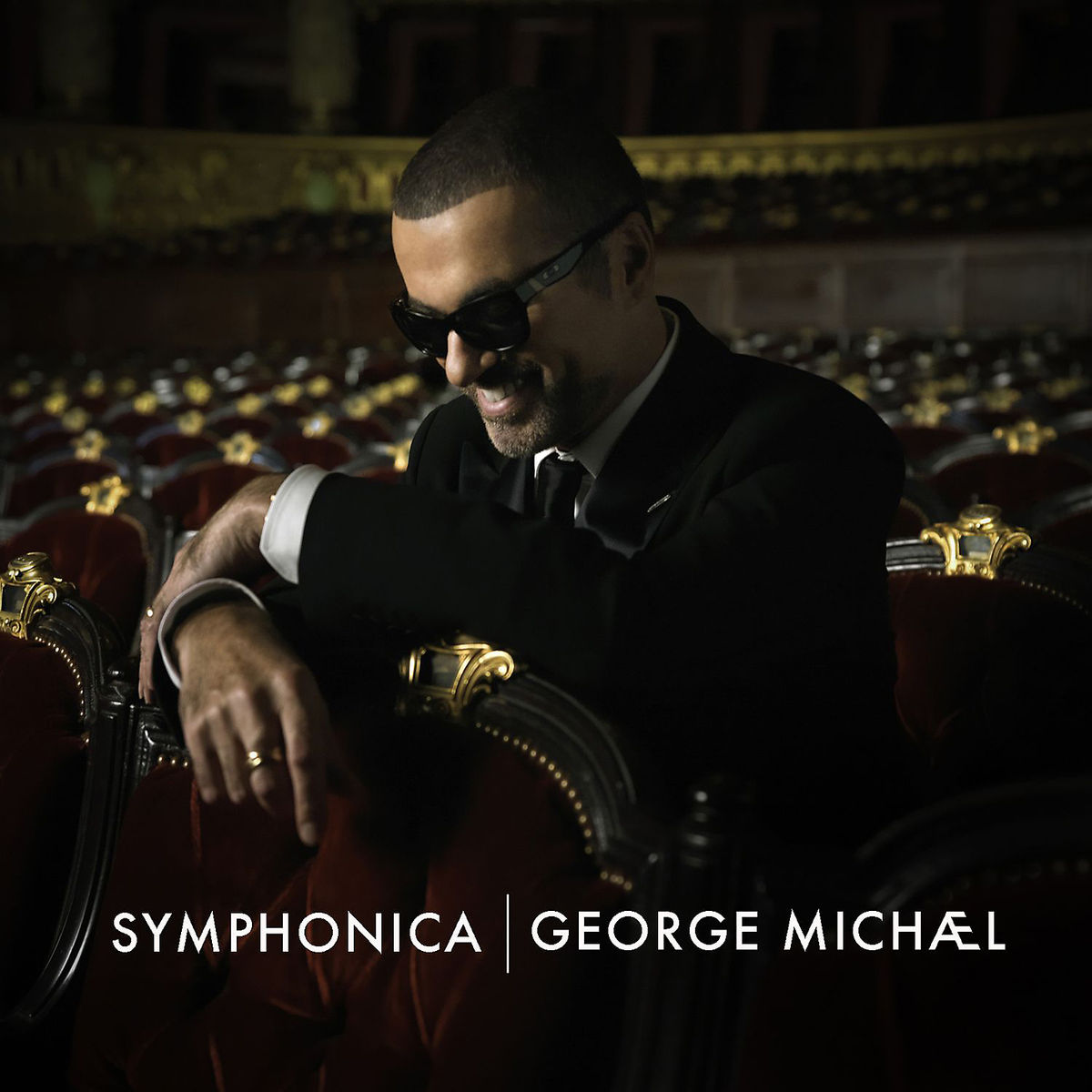 George Michael im Anzug und Sonnenbrille, im Theater sitzend. Das Wort 'Symphonica' und sein Name stehen unten.
