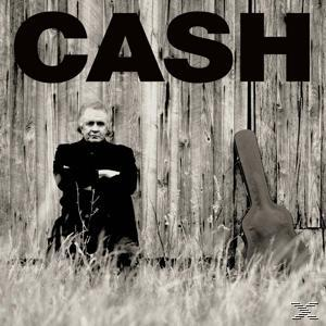 Ein Schwarzweißfoto von Johnny Cash. Er lehnt mit einem Gitarrenkoffer an einem Holzzaun.