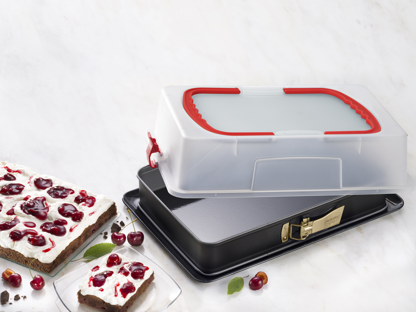 Kuchen in einem Behälter und zwei Kuchenstücke auf Marmor. Kirschen sind verstreut.