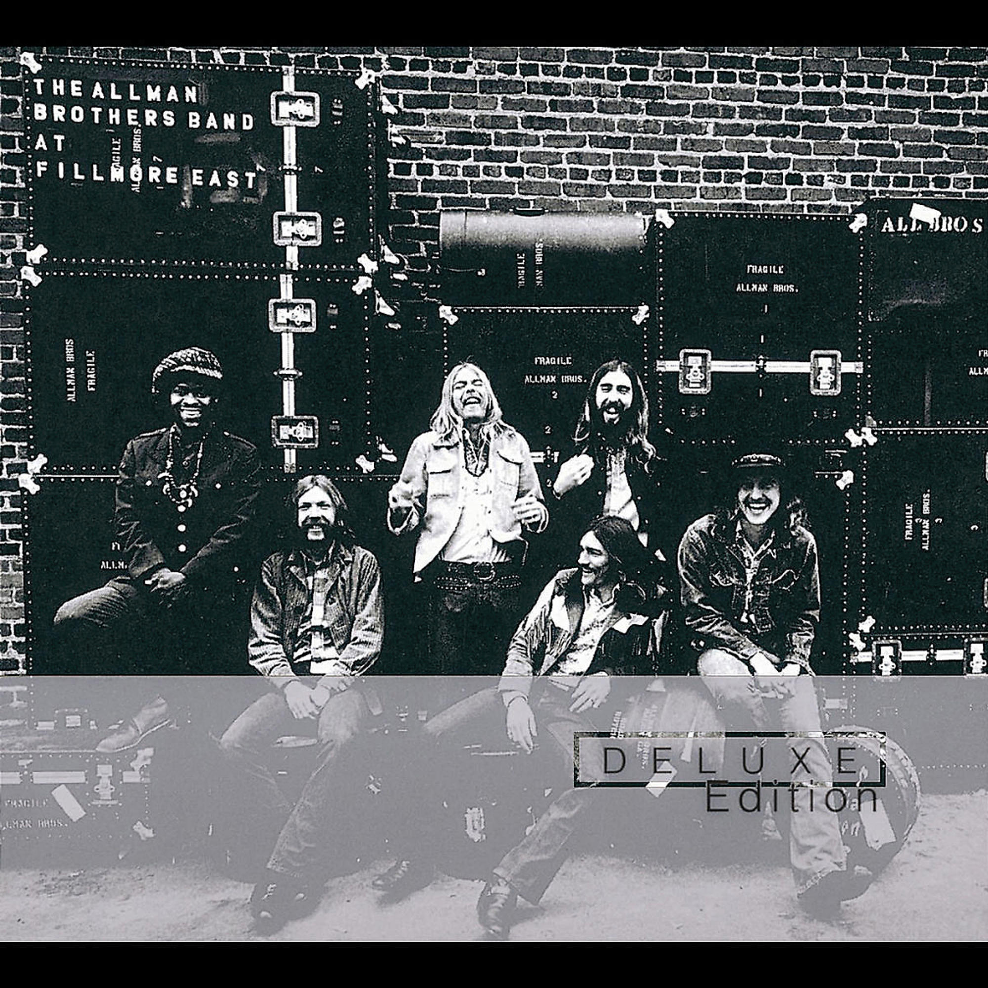Albumcover mit einer Band. Gruppe von sieben Männern vor Backsteinmauern mit Versandcontainern.