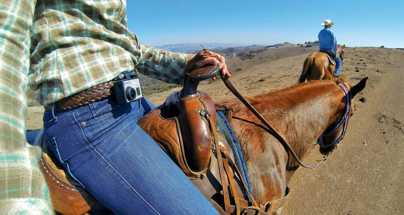 Twee mensen te paard. Eén staat op de voorgrond met een geruit overhemd en houdt een camera vast.