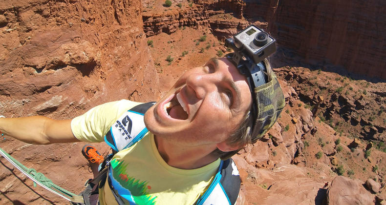 Man op een klif, met een GoPro op een helm, kijkt omhoog en schreeuwt, met een rode rotsachtergrond.