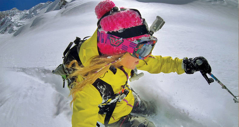 Een skiër in het geel skiet een besneeuwde berg af. De skiër draagt een roze helm en een bril.