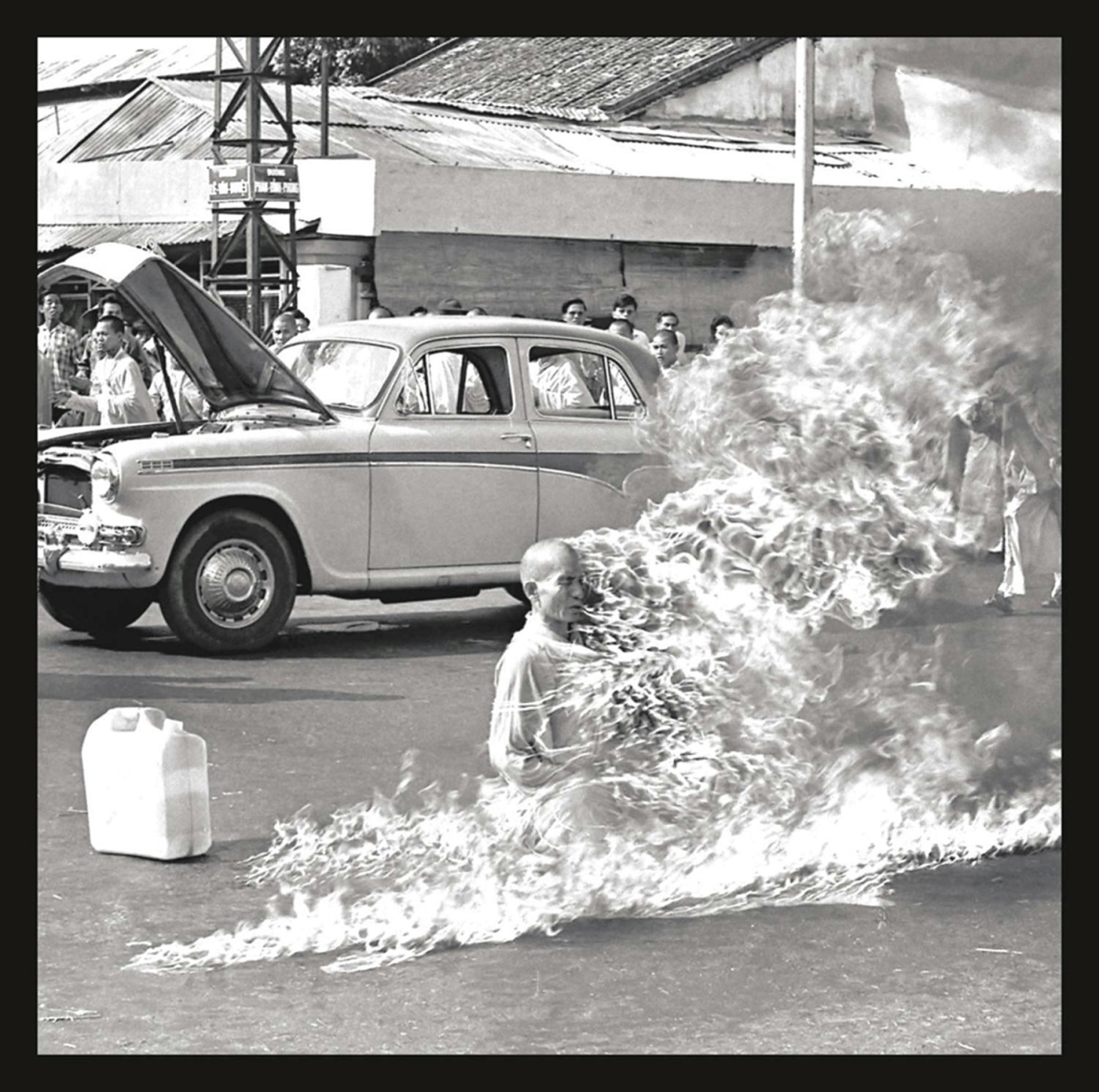 Schwarz-Weiß-Foto. Ein brennender Mann vor einem Auto. Leute schauen zu.