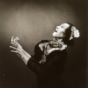 Monochrome Foto einer Frau in traditioneller Kleidung, die nach oben schaut und die Hände hebt. Dunkler Hintergrund.