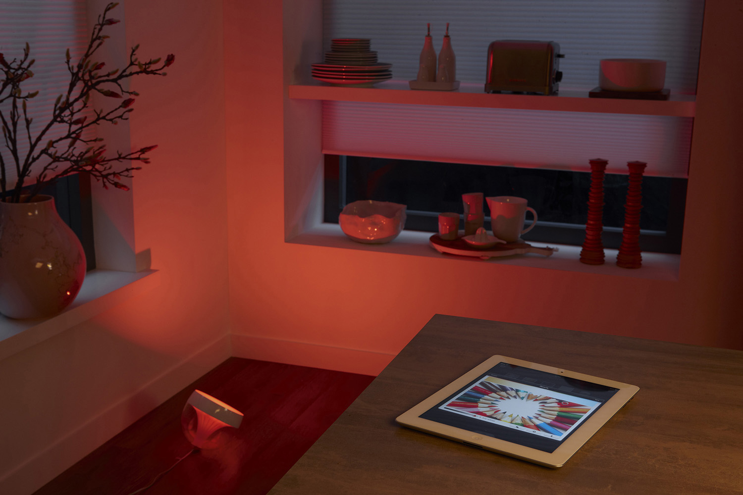 Ein Raum mit rot beleuchteter Wand und Tisch mit Tablet, das Farbpalette zeigt. Vase und Fenster.
