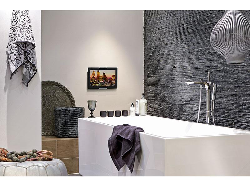 Badezimmer mit Fernseher, Badewanne, Handtüchern und Dekorationsartikeln. Dunkle Steinwand und eine große Lampe im Hintergrund.