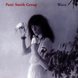 Patti Smith hält zwei weiße Tauben. Sie trägt ein weißes Kleid, hat dunkles Haar und steht vor einer hellen Wand.
