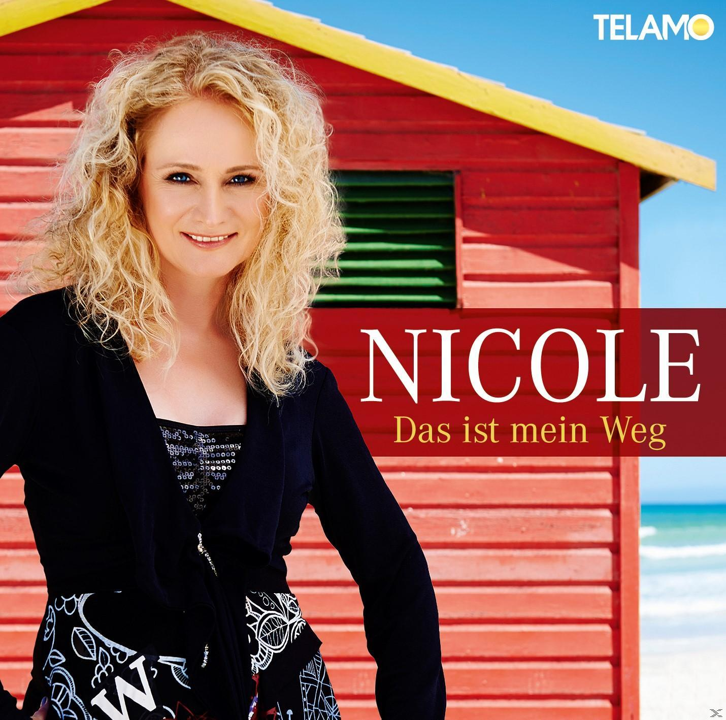 Frau mit blonden Locken steht vor einem roten Gebäude mit Text.