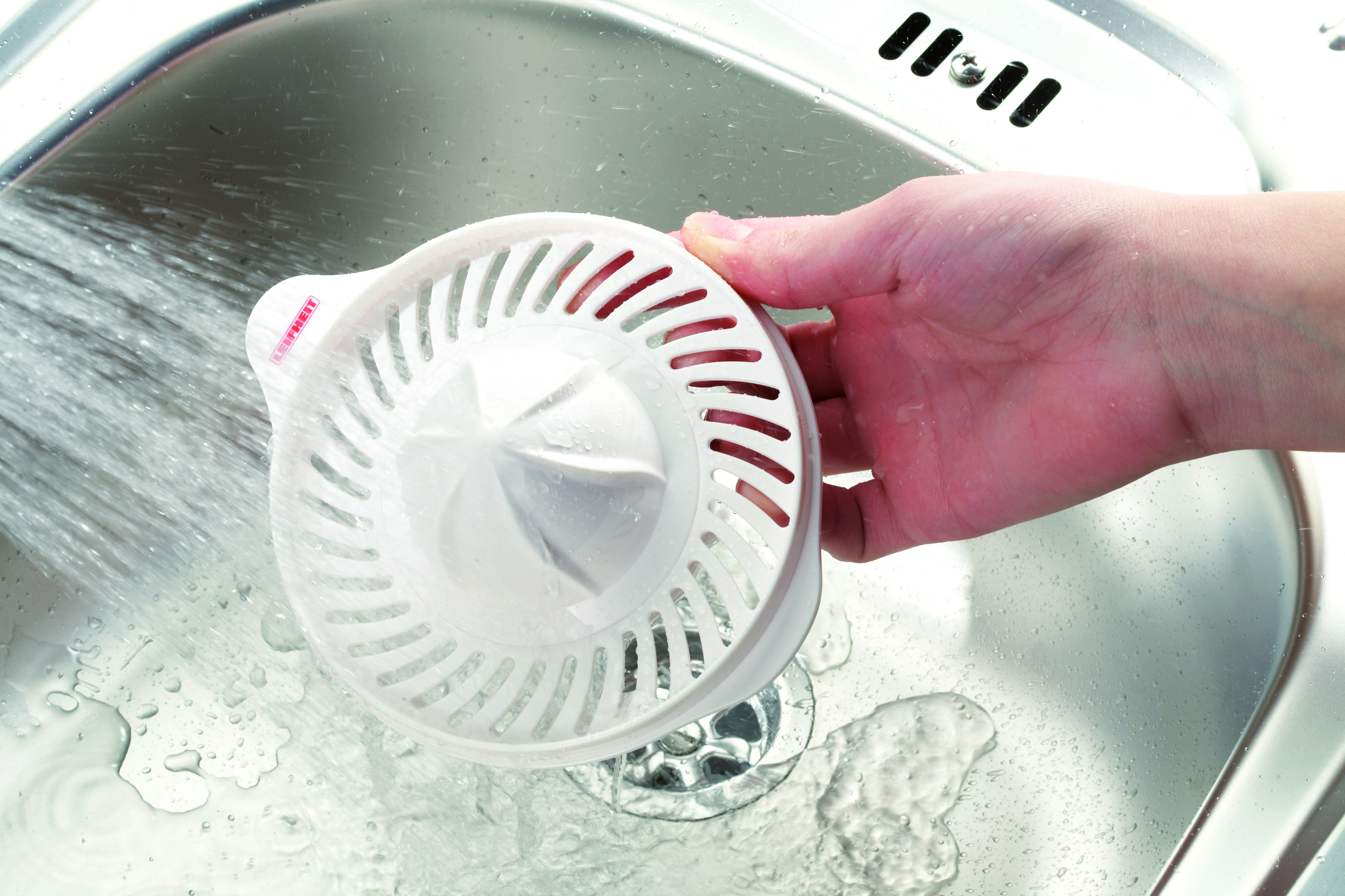 Eine Hand wäscht einen weißen Saftpressen-Sieb unter fließendem Wasser im Waschbecken.