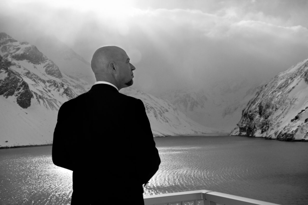 Schwarzweißfoto eines Mannes im Anzug, der eine schneebedeckte Bergkette und einen See betrachtet.