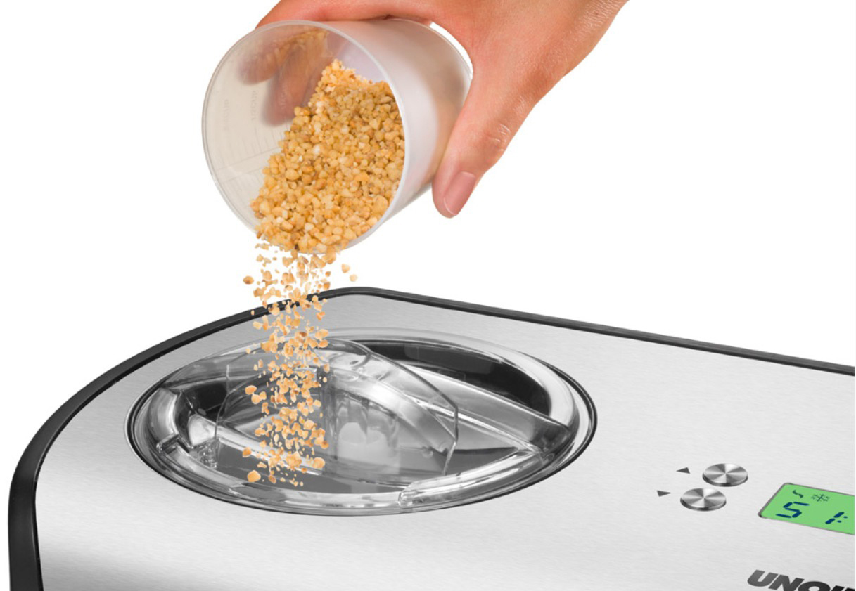 Eine Hand, die kleine braune Granulate aus einem Messbecher in eine silberne Küchenmaschine gießt.