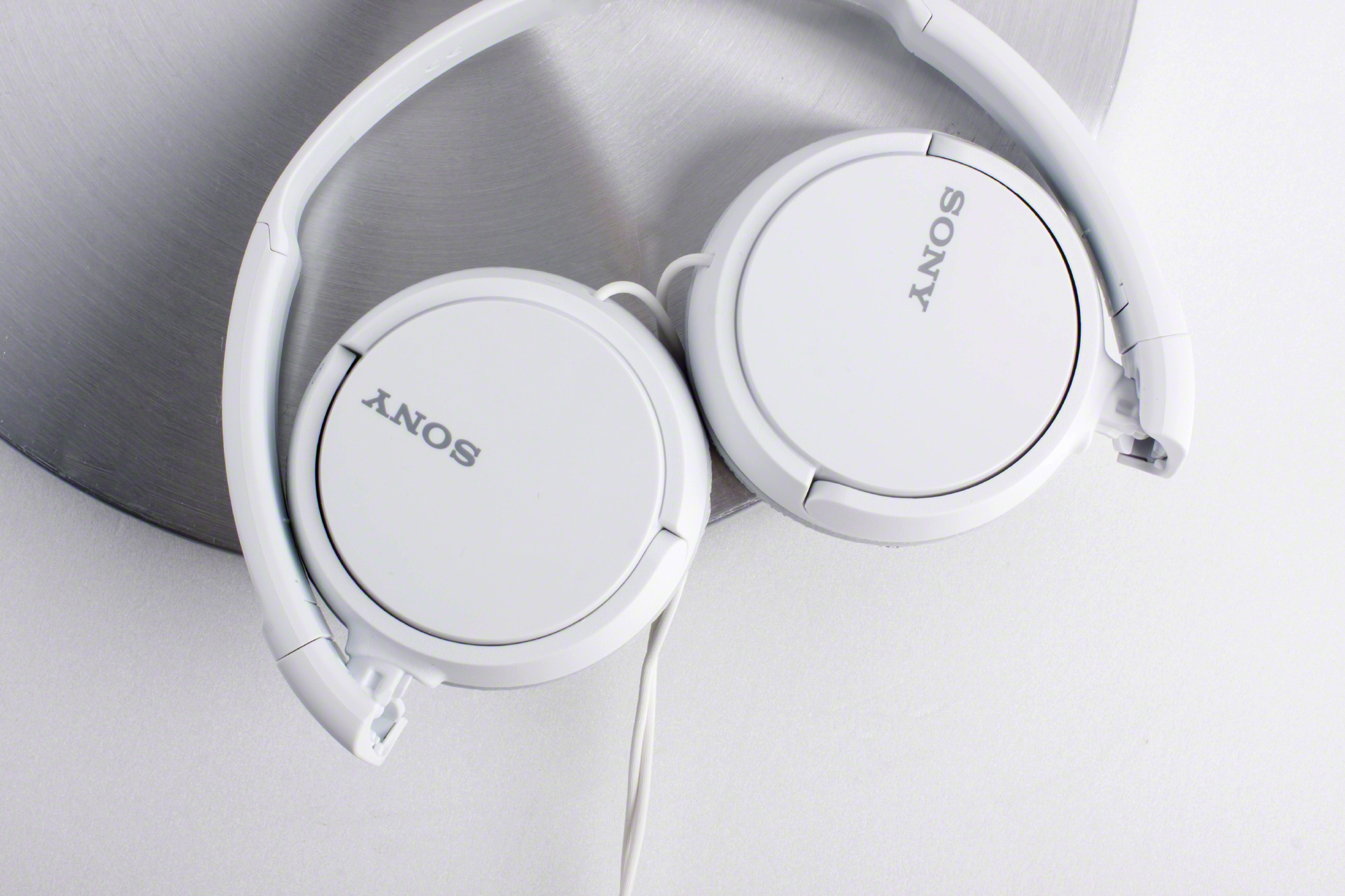 Faltbare weiße Sony-Kopfhörer, teilweise geöffnet, auf einer kreisförmigen Metalloberfläche platziert. Einfach und sauber.