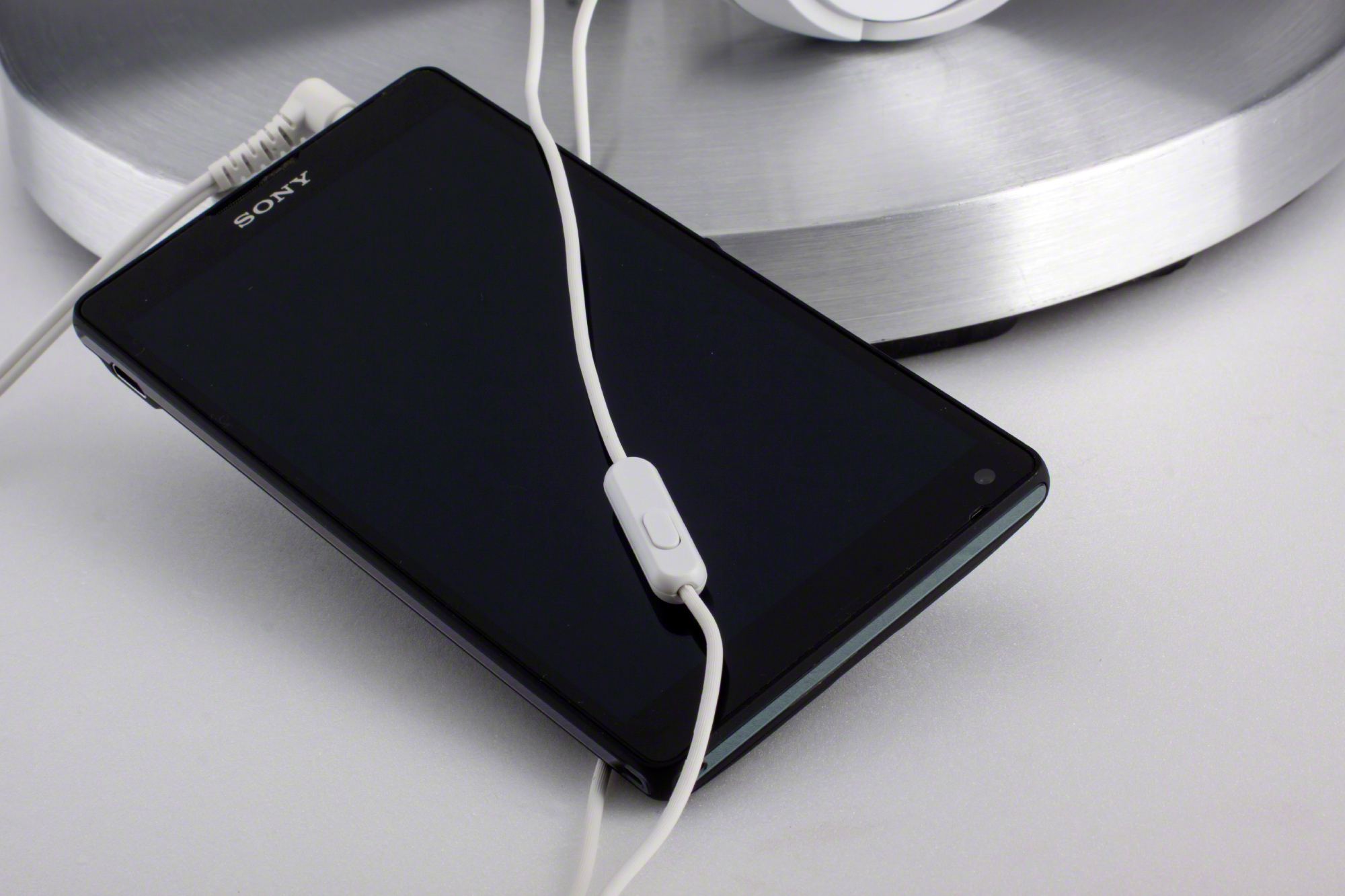Schwarzes Sony-Telefon mit weißen Kopfhörern, die eingesteckt sind, auf einer silbernen Oberfläche.