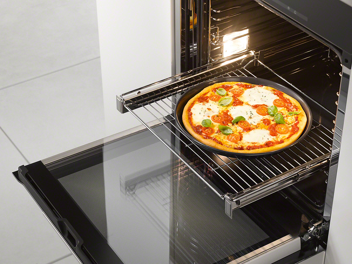 Ofen mit Pizza darin. Die Pizza hat Tomaten, Basilikum und Käse. Die Ofentür ist offen und das Licht ist an.