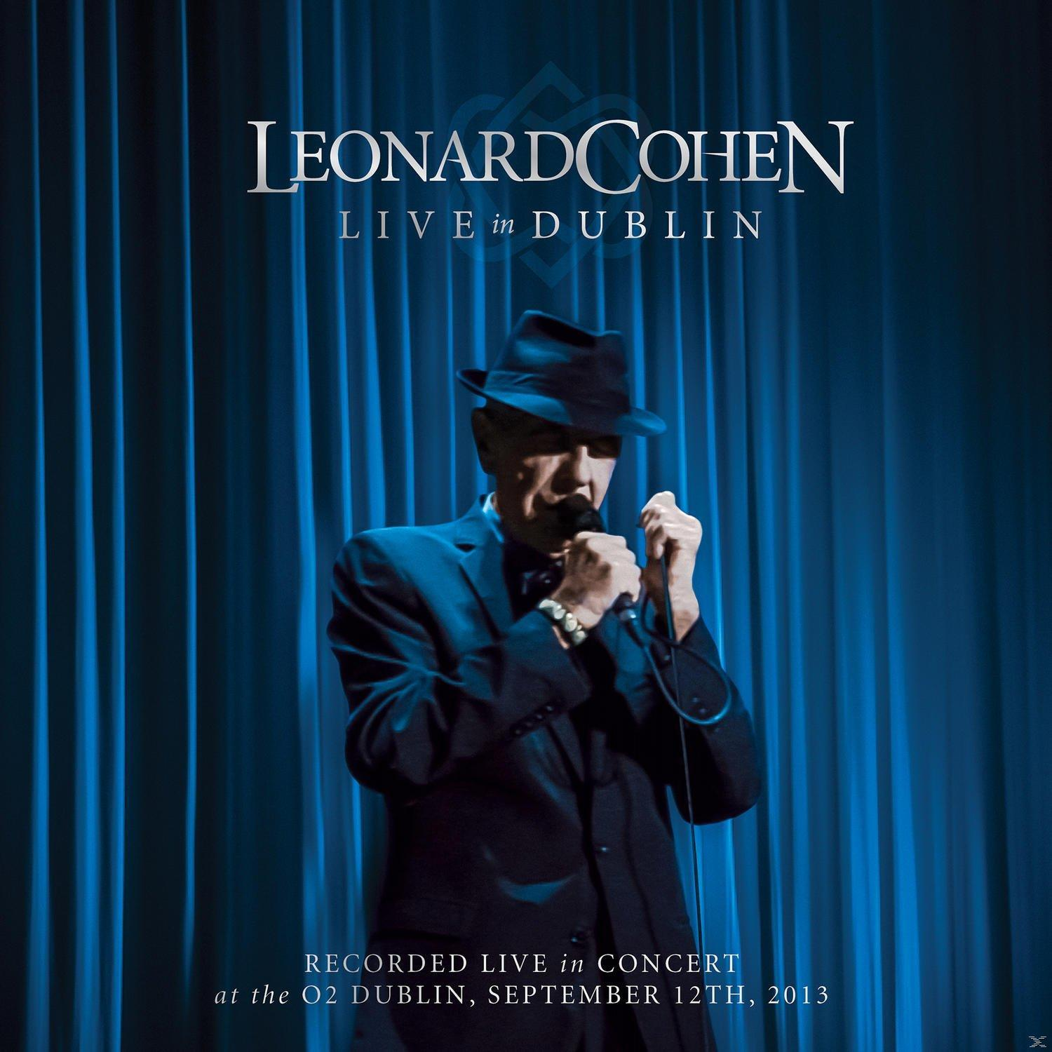 Leonard Cohen op het podium, zingend, in pak en hoed, voor een blauw gordijn.