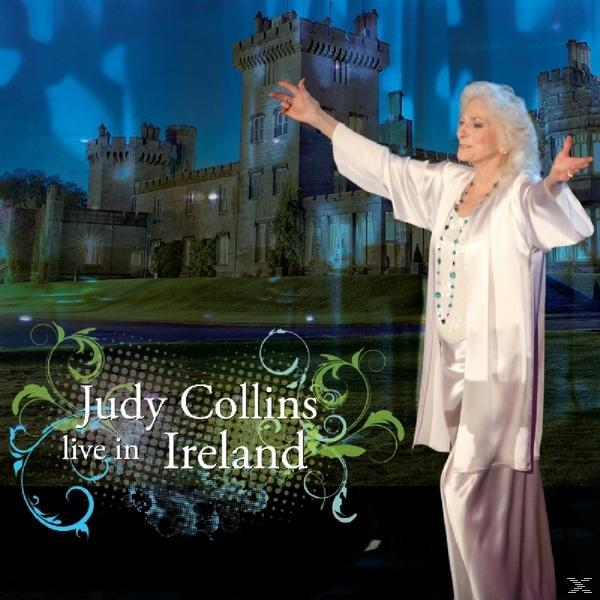 Judy Collins tritt auf einer Bühne vor einer Schlosskulisse auf, Arme ausgestreckt, in Weiß gekleidet.