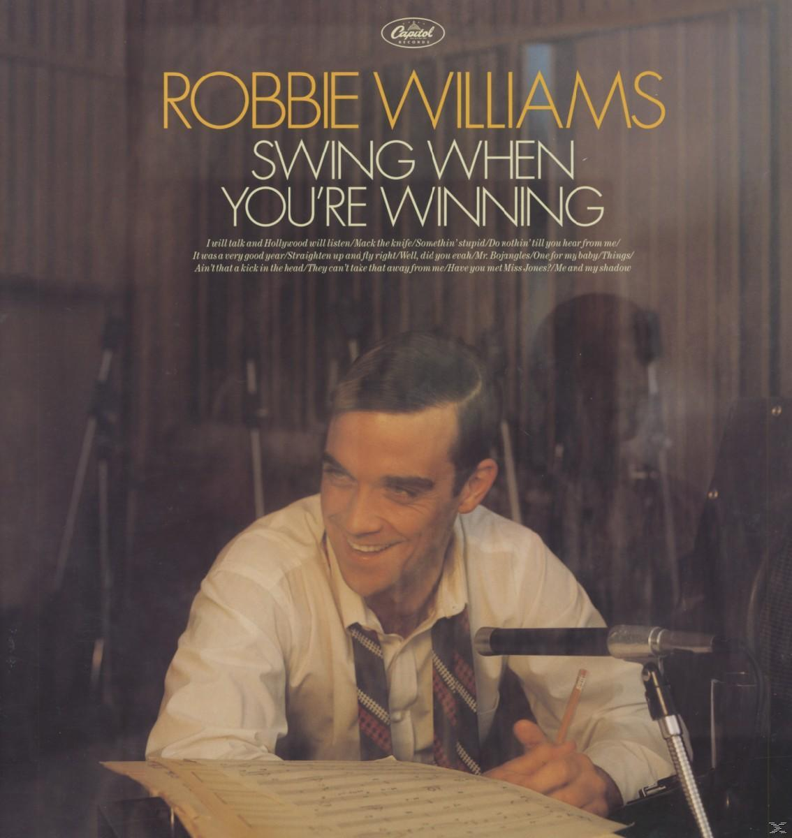 Albumcover mit Robbie Williams lächelnd, Bleistift haltend, Noten. Goldener Text.