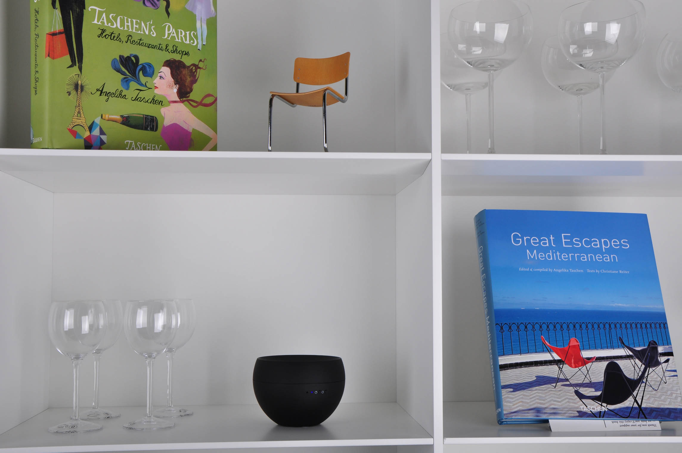 Weiße Regale mit Büchern, Gläsern und einem schwarzen Lautsprecher. Bücher: Taschen's Paris, Great Escapes.